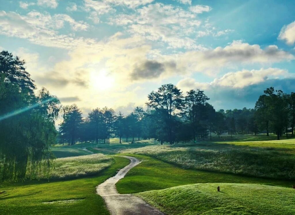 Sun pierces through the light clouds to illuminate the tee box, cart path, and bordering tall grass.